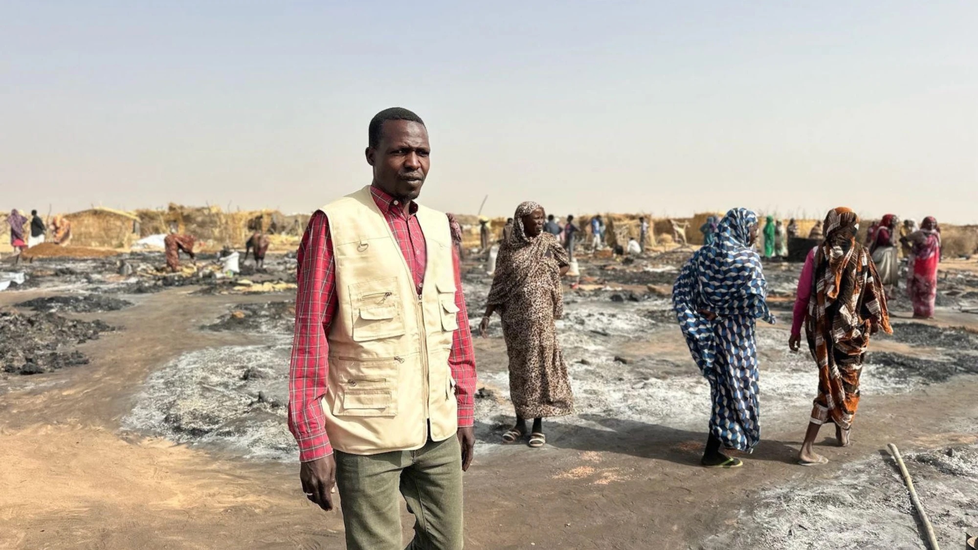 An aid worker in a beige vest stands on scorched ground where a settlement has burned, women walking among the ashes behind him