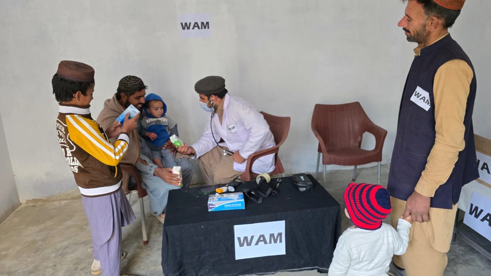 A clinician in a white coat and mask listens to a child held by his father, in a bare room with WAM signs on the wall and table