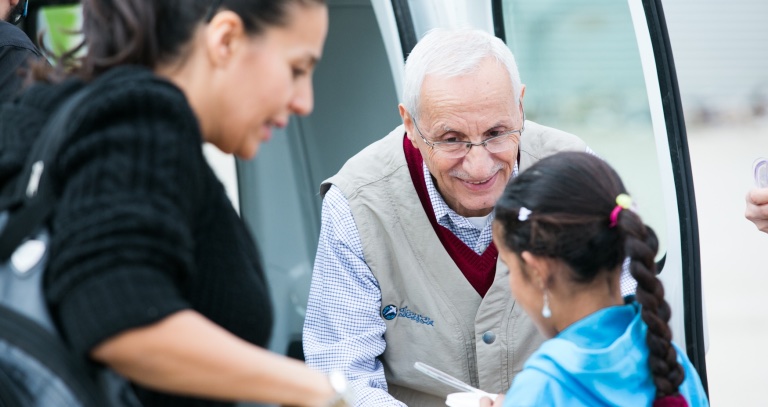 Dr. Jean Bouchebel smiling as he hands something to a young girl beside a woman at the open door of a vehicle