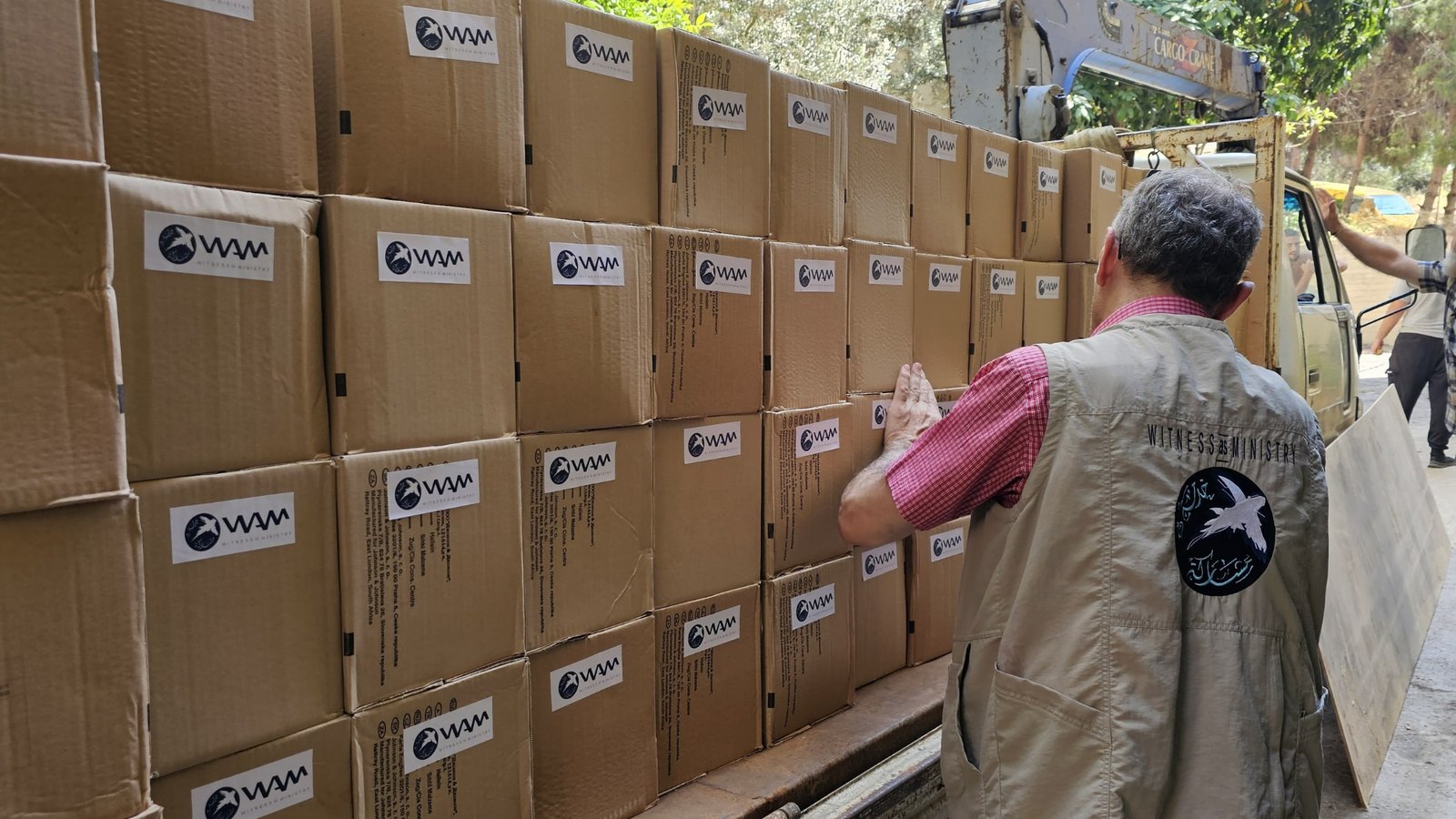 A WAM worker in a Witness as Ministry vest steadies a wall of stacked cartons, each marked with the WAM logo, beside a lorry