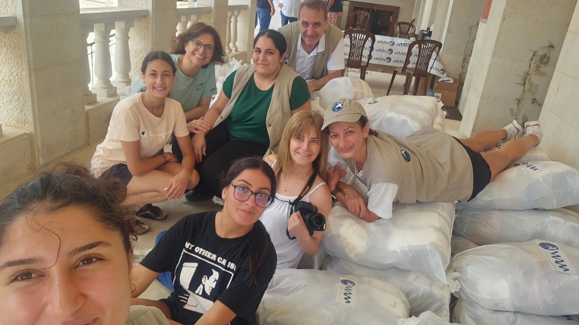 A WAM team of volunteers, smiling for a selfie among stacked sacks of relief parcels on a stone-arched terrace